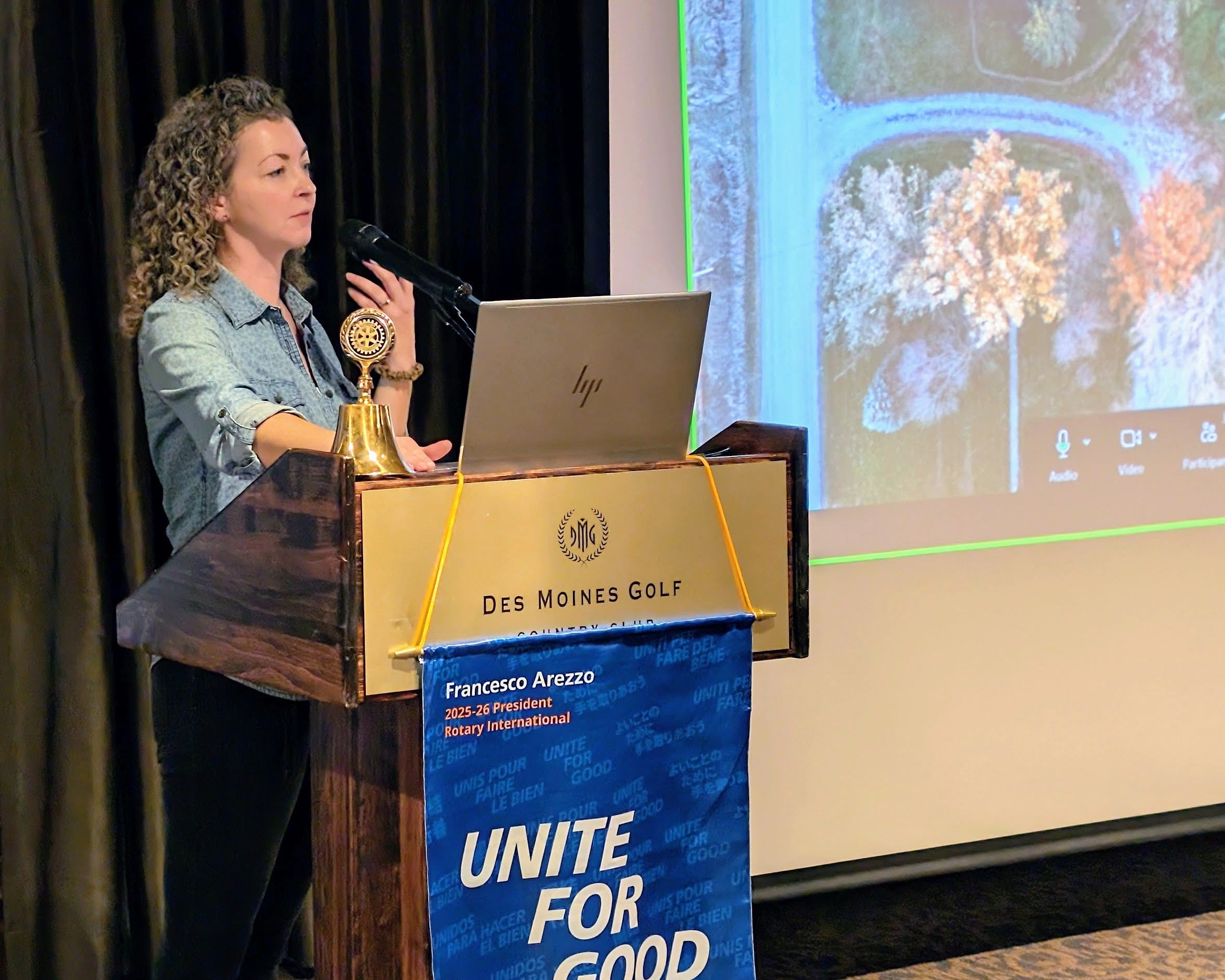 Woman at a podium with a laptop, bell, and Des Moines Golf banner in a conference setting.