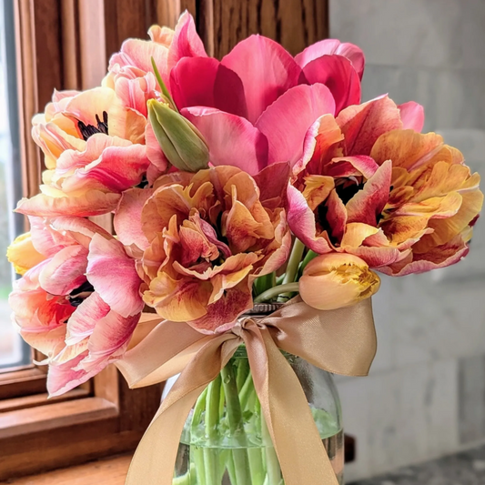 Bouquet of pink, orange, and yellow tulips in a clear vase with a beige ribbon.